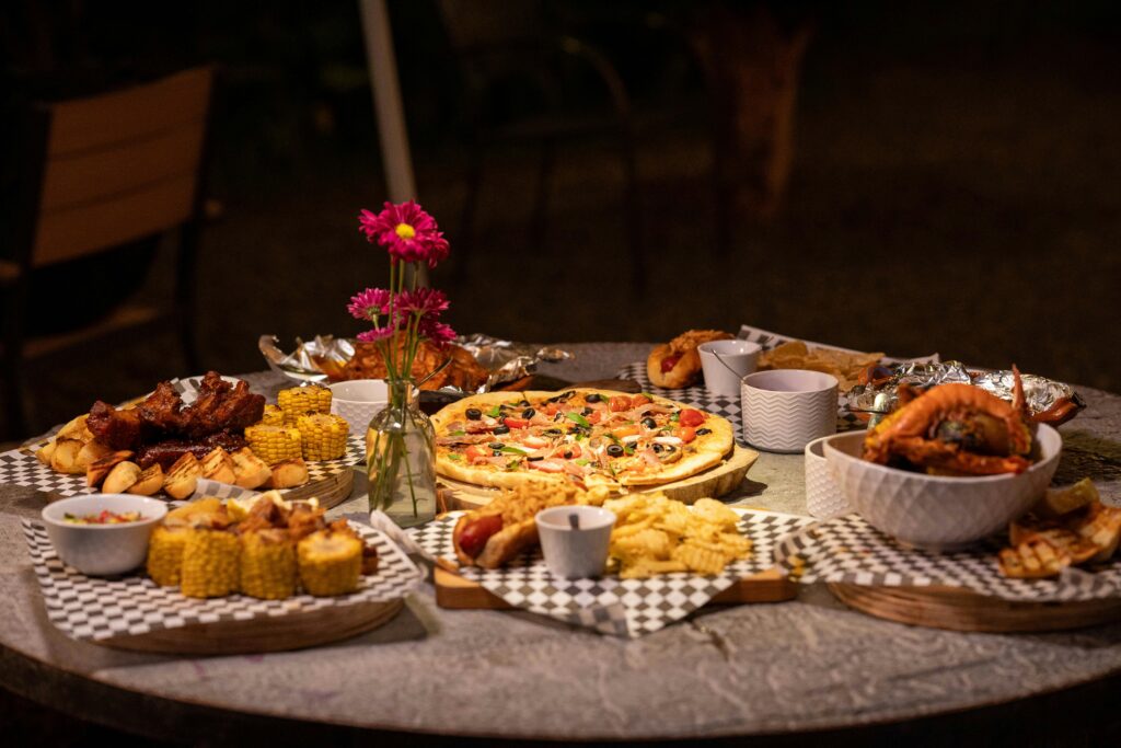 pexels-photo-35896428-35896428 A diverse dinner spread with pizza, corn, ribs, and snacks on an outdoor table setting.